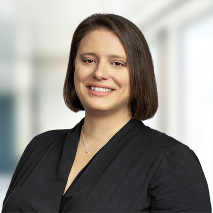 A woman with straight brown hair, wearing a black top and a necklace, smiles at the camera in a brightly lit corporate law office.