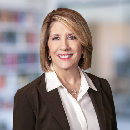 A woman with shoulder-length light brown hair, wearing a white collared shirt and dark blazer, smiles in an office setting with blurred shelves in the background, reflecting the professional atmosphere of Chicago lawyers.