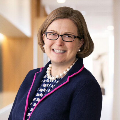 A woman wearing glasses, a navy blazer with pink trim, a patterned blouse, and a pearl necklace smiles in the brightly lit hallway of law offices specializing in intellectual property law.