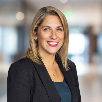 A woman with blonde hair wearing a black blazer and teal top smiles in a blurred office setting, reflecting the professionalism often seen at law offices specializing in intellectual property law.