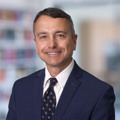 A man in a navy suit and patterned tie smiles at the camera in a professional law offices setting with blurred bookshelves in the background, portraying the confident demeanor of lawyers in Chicago.