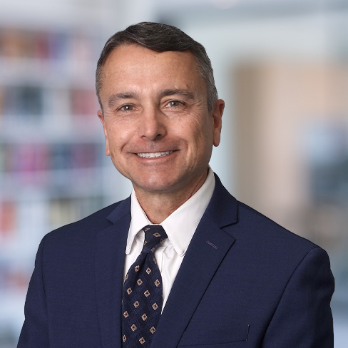 A man in a navy suit and patterned tie smiles at the camera in a professional law offices setting with blurred bookshelves in the background, portraying the confident demeanor of lawyers in Chicago.