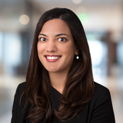 A woman with long dark hair, wearing a black top, smiles at the camera in a blurred corporate law office background.