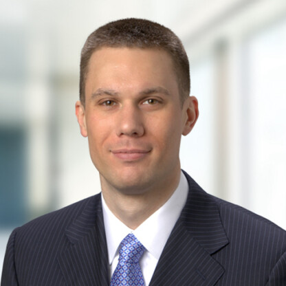 A man in a dark pinstripe suit and blue patterned tie poses for a formal headshot against a blurred indoor background, reflecting his professionalism in law offices and expertise in intellectual property law.