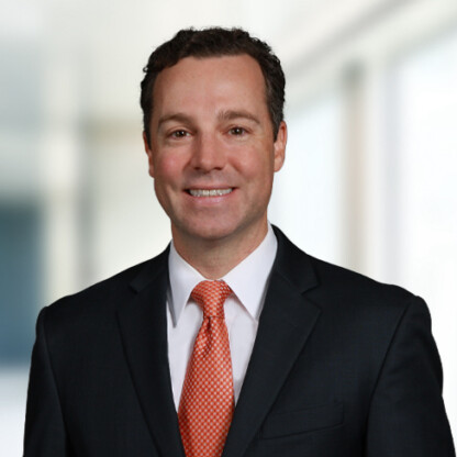 A man in a dark suit and orange tie smiles at the camera, standing in front of a blurred indoor office background, reflecting the professional atmosphere typical of law offices specializing in intellectual property law.