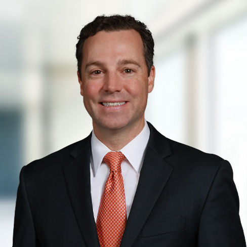 A man in a dark suit and orange tie smiles at the camera, standing in front of a blurred indoor office background, reflecting the professional atmosphere typical of law offices specializing in intellectual property law.