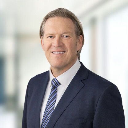 A man in a navy suit with a striped tie smiles at the camera, standing against a blurred indoor setting of a corporate law office.