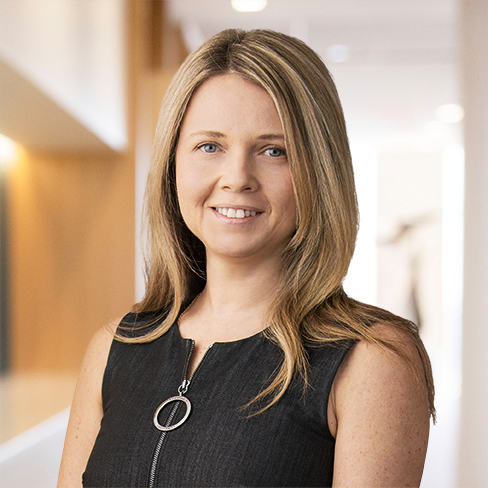 A woman with long blonde hair wearing a sleeveless black dress stands in a bright, modern corporate law office, smiling at the camera.