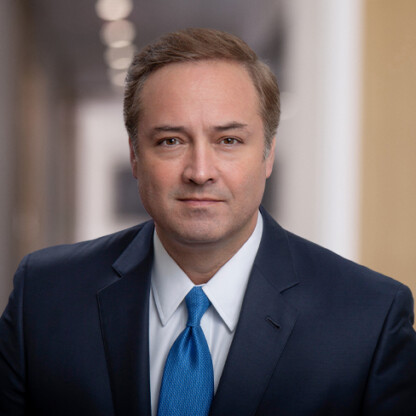 A man in a suit and blue tie poses for a formal portrait in the hallway of law offices, with blurred lights in the background, reflecting his expertise in litigation support.