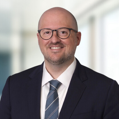 A man wearing glasses, a dark suit, a white shirt, and a striped tie smiles confidently in front of a blurred indoor background, embodying the professionalism found at top law offices specializing in intellectual property law.