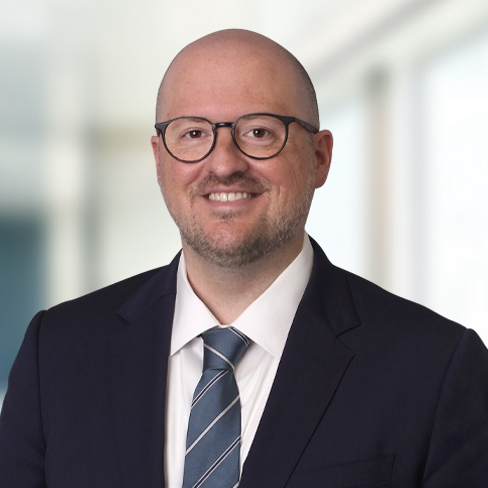 A man wearing glasses, a dark suit, a white shirt, and a striped tie smiles confidently in front of a blurred indoor background, embodying the professionalism found at top law offices specializing in intellectual property law.