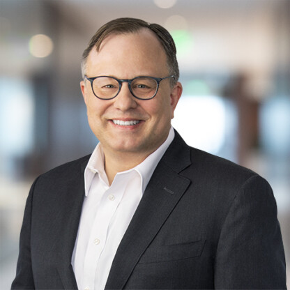 A man wearing glasses, a white shirt, and a dark suit jacket smiles while standing in a blurred indoor corporate law office setting.