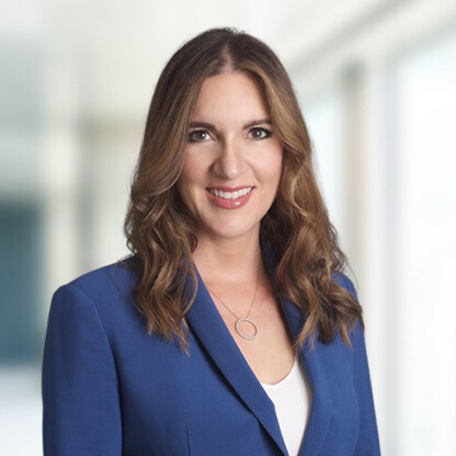 Woman with long brown hair wearing a blue blazer and white top, standing in a brightly lit corporate law office, facing the camera and smiling.