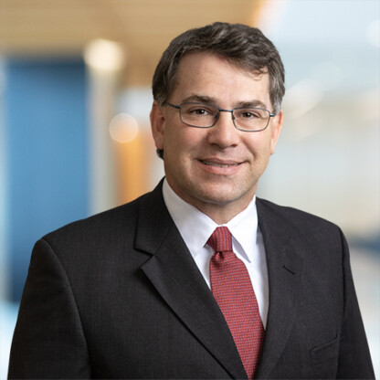 A man in a dark suit, white shirt, and red tie, wearing glasses, stands in front of a blurred corporate law office background.