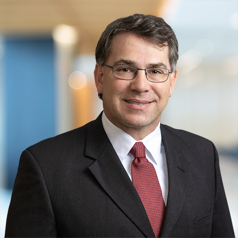A man in a dark suit, white shirt, and red tie, wearing glasses, stands in front of a blurred corporate law office background.