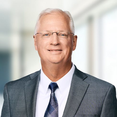 An older man with short white hair, glasses, and a gray suit with a blue tie stands in a brightly lit corporate law office environment.