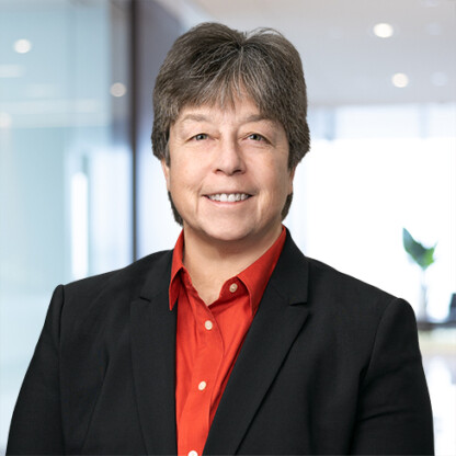 A person with short gray hair wearing a black blazer and red shirt stands in a modern corporate law office, smiling at the camera.