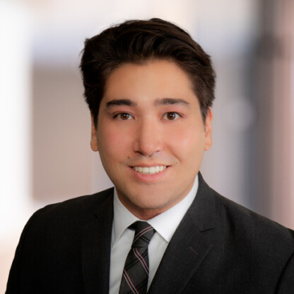 A man with dark hair wearing a dark suit, white shirt, and striped tie, smiling at the camera against a blurred background in a corporate law office of Chicago lawyers.