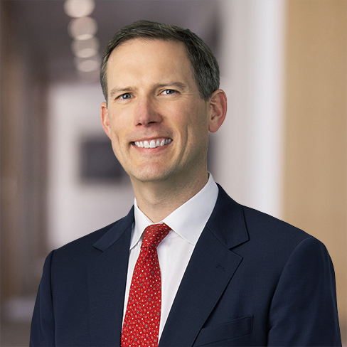 A man in a navy suit and red tie smiles at the camera, standing in a blurred corporate law office hallway background.