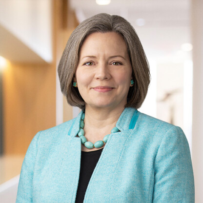 A woman with short gray hair, wearing a turquoise blazer, matching necklace, and earrings, stands indoors in a well-lit, modern hallway at a corporate law office.