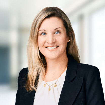 A woman with long blonde hair, wearing a dark blazer over a light blouse and a gold necklace, smiles at the camera in a bright, blurred corporate law office setting.