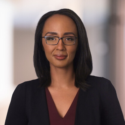 A woman with straight shoulder-length dark hair, glasses, and a burgundy top under a black blazer stands against a blurred indoor background, capturing the professional atmosphere of a corporate law office.