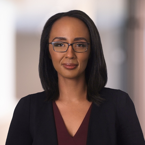 A woman with straight shoulder-length dark hair, glasses, and a burgundy top under a black blazer stands against a blurred indoor background, capturing the professional atmosphere of a corporate law office.
