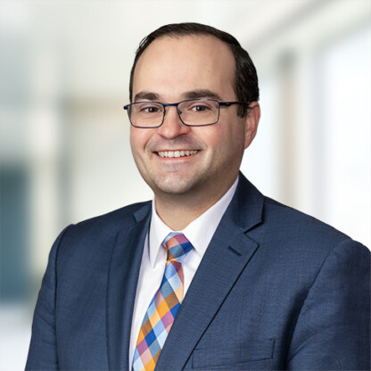 A man in a blue suit, patterned tie, and glasses smiles at the camera with a blurred corporate law office background.