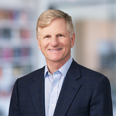 A middle-aged man with short blond hair, wearing a blue suit jacket and light blue shirt, smiles at the camera in a corporate law office.
