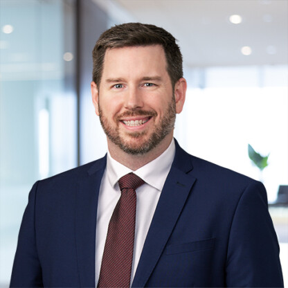 A man with short brown hair and a beard, wearing a navy suit, white shirt, and red tie, stands indoors in a modern corporate law office.