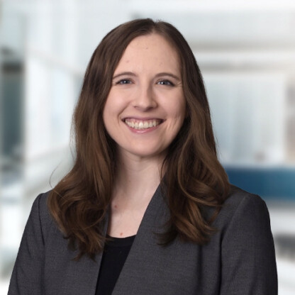 Woman with long brown hair, wearing a grey blazer over a black top, smiling at the camera in a bright, blurred corporate law office, representing the professionalism of Chicago lawyers.