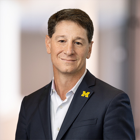 A man in a navy suit and white shirt wears a University of Michigan "M" pin on his lapel, standing in an indoor setting with a blurred background, likely within a corporate law office.