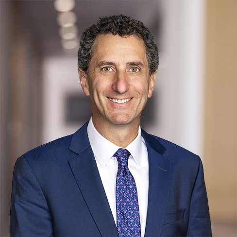 A man with curly dark hair in a blue suit and patterned tie smiles at the camera in the well-lit hallway of a corporate law office.