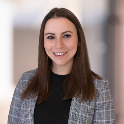 A woman with straight brown hair wearing a gray plaid blazer and black top, smiling at the camera in a softly blurred indoor setting, exemplifying professionalism often seen among Chicago lawyers.