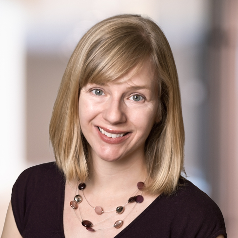 A woman with straight blonde hair and bangs smiles at the camera. She is wearing a dark top and a beaded necklace, embodying the professionalism seen in lawyers in Chicago. The background is blurred.
