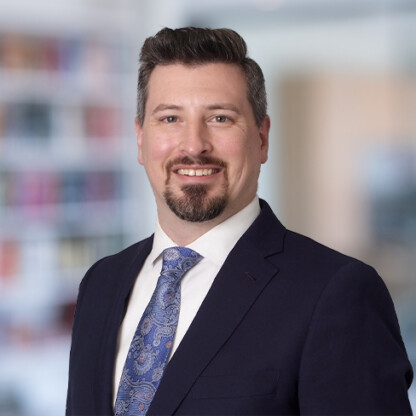 A man in a navy suit, white shirt, and patterned blue tie, smiling, stands in front of a blurred corporate law office background with shelves and books.