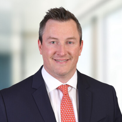 A man in a dark suit, white shirt, and patterned red tie is smiling in front of a blurred law offices background.