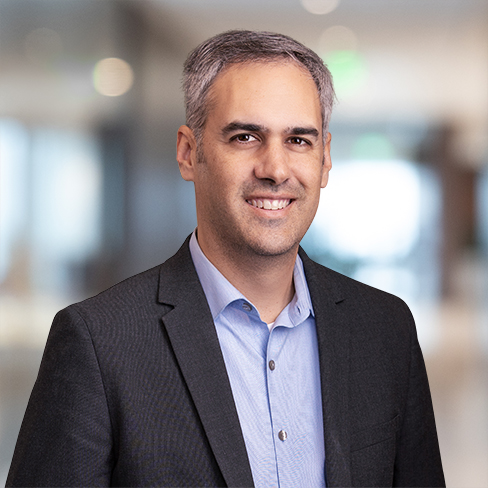 A man with short gray hair wearing a dark blazer and light blue shirt smiles, standing in a blurred indoor office setting—reflecting the professionalism of lawyers in Chicago specializing in litigation support.