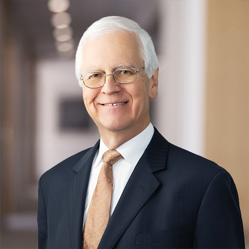An older man with white hair and glasses, wearing a dark suit, white shirt, and beige tie, stands in a blurred hallway of a corporate law office offering litigation support.