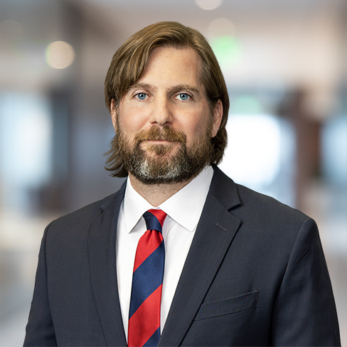 A man with shoulder-length brown hair and a beard, wearing a dark suit, white shirt, and red-and-blue striped tie, stands in the blurred backdrop of a bustling corporate law office.