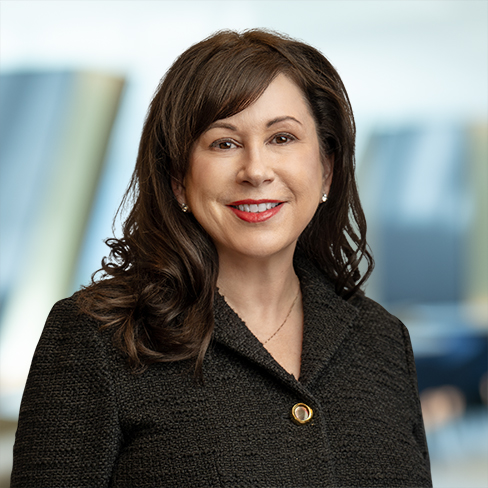 A woman with long brown hair wearing a dark textured blazer and a gold pin smiles at the camera, set against a blurred law offices background.