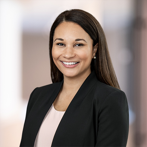 A woman with long brown hair, wearing a black blazer and light pink top, smiles at the camera against a blurred office background, reflecting the professionalism of Chicago lawyers in leading law offices.