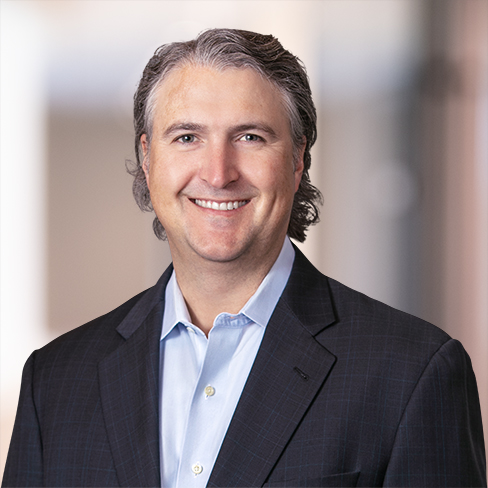 A man with graying hair wearing a dark suit jacket and light blue shirt, smiling at the camera with a blurred law offices background, reflecting the professionalism of lawyers in Chicago.