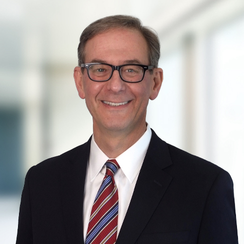 A man in a suit, tie, and glasses smiles at the camera in a brightly lit, blurred corporate law office—perfectly capturing the professionalism of lawyers in Chicago.