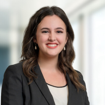 A woman with long brown hair, wearing a black blazer, cream blouse, and pearl earrings, smiles at the camera in a bright corporate law office with a blurred indoor setting.