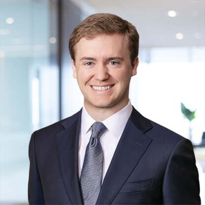 A young man in a dark suit and tie smiles in a modern corporate law office setting, with glass walls and bright lighting.