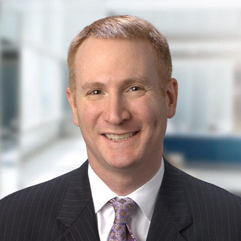 Smiling man in a dark pinstripe suit and patterned tie, with short light brown hair, posing in a bright, blurred corporate law office—ideal for showcasing experienced lawyers in Chicago.