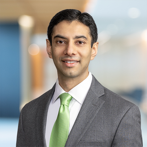 A man in a gray suit, white shirt, and light green tie stands indoors with a blurred corporate law office background.
