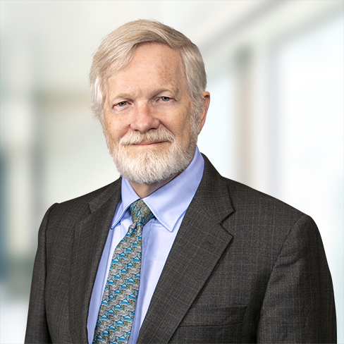An older man with gray hair and a beard, dressed in a dark suit, blue shirt, and patterned tie, stands confidently in front of a blurred indoor background, representing one of the top Chicago lawyers specializing in intellectual property law.