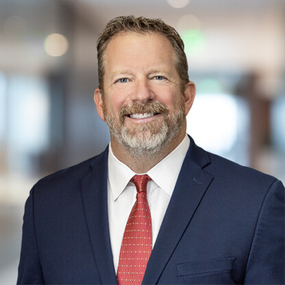A middle-aged man in a navy suit, white shirt, and red tie smiles at the camera in a blurred corporate law office, suggesting expertise in intellectual property law.
